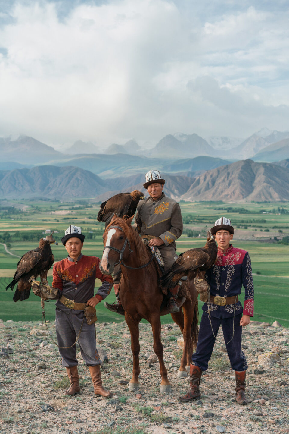 Three traditional eagle hunters in Kyrgyzstan with golden eagles and a horse against a mountain landscape.